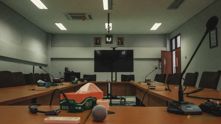 Empty conference room with long tables, microphones, a large display screen, and a hanging ceiling microphone ready for a meeting or hybrid session.